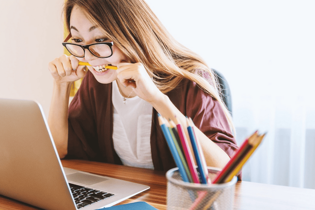A woman with glasses sitting at a desk with pencils in a jar, looking at her laptop whilst holding a pencil in her mouth